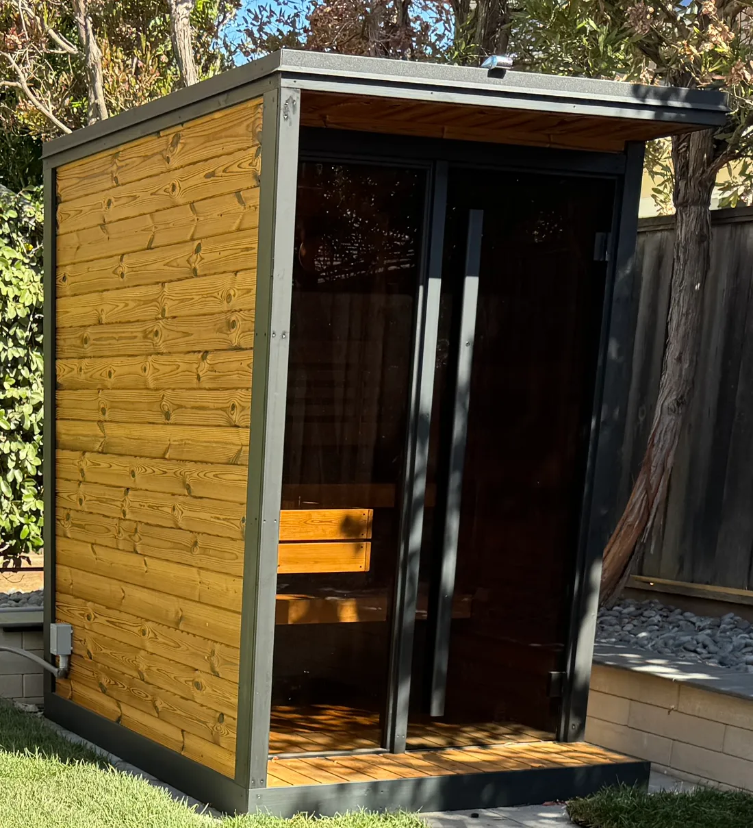 An outdoor prefab sauna with dark glass doors on a gravel pad, Foster City